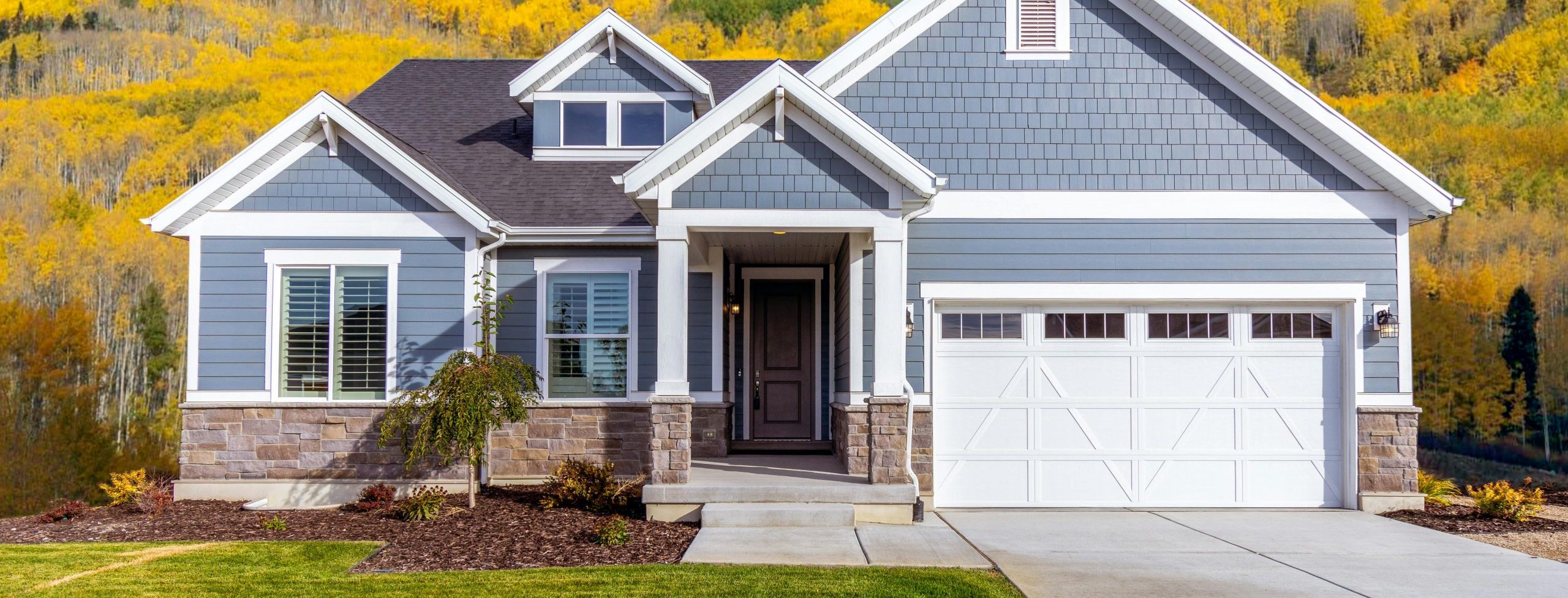 Front view of a blue house in a mountain neighborhood, surrounded by trees with autumn-colored leaves, used to indicate now is a good time to sell your home.