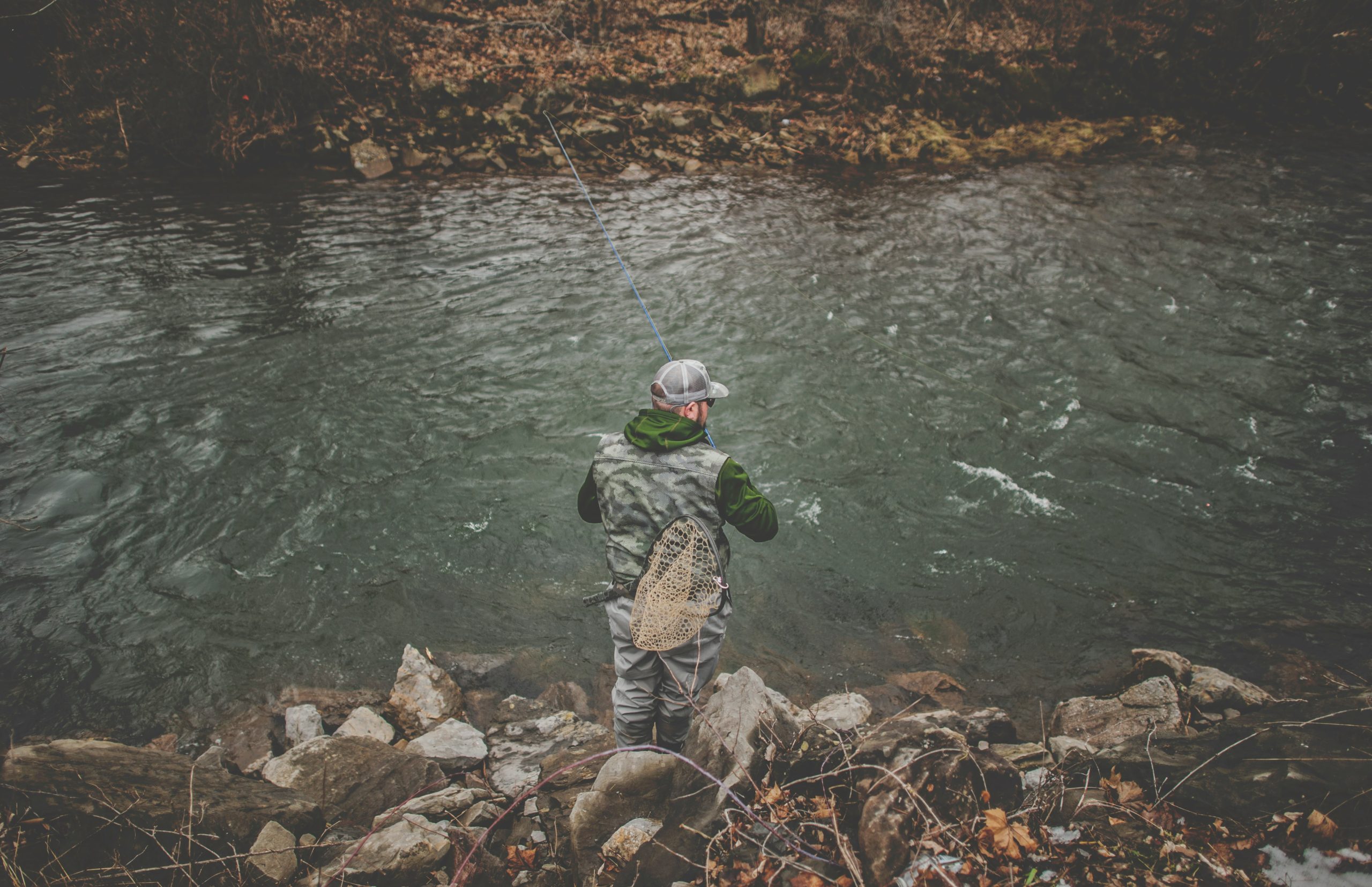 Man fishing by a riverfront and standing on the rocks, facing away from the camera. 