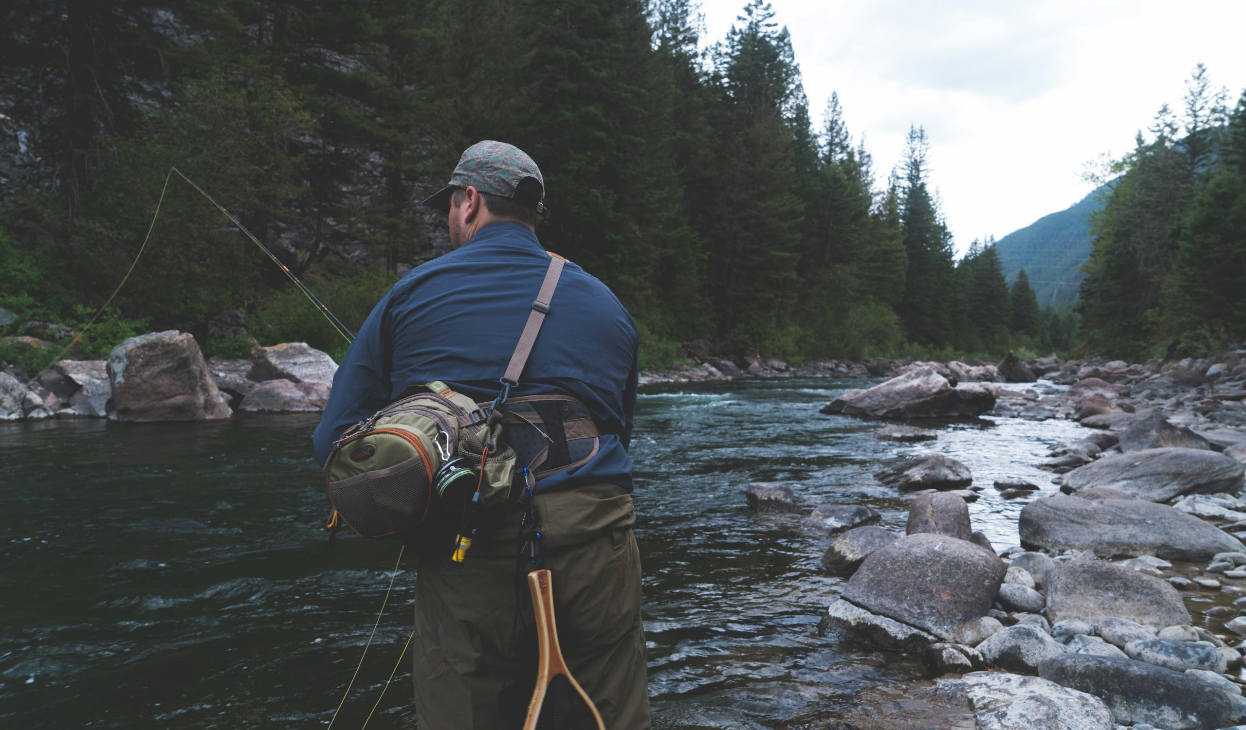 Man fishing in a rocky creek with trees surrounding him on an overcast day. 
