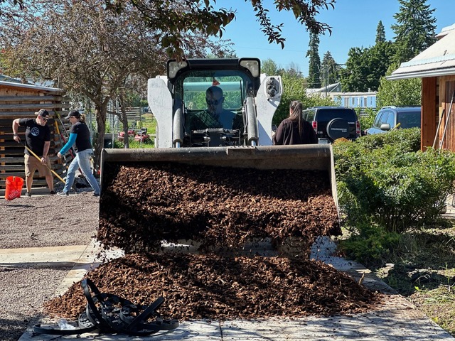 A bulldozer full of woodchips is placed onto the ground while volunteers work in the background.