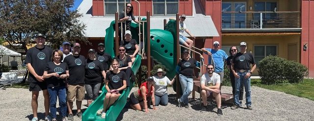 A group of volunteers gathers in and around a large piece of playground equipment and smiles.